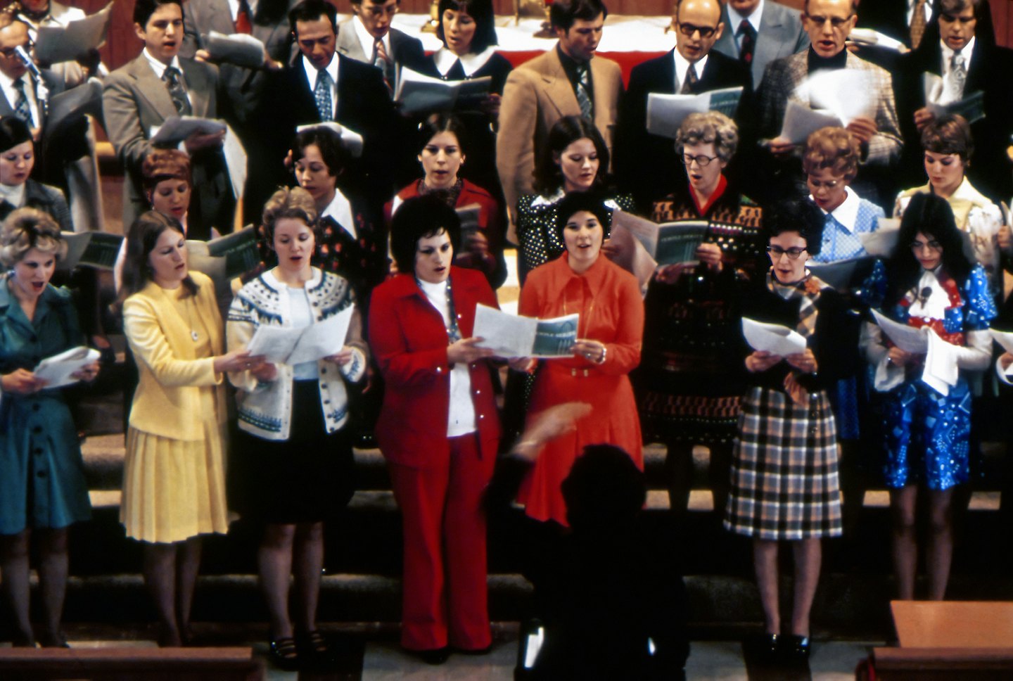 Een koor zingt van bladmuziek op verhoogde podia onder leiding van een dirigent.